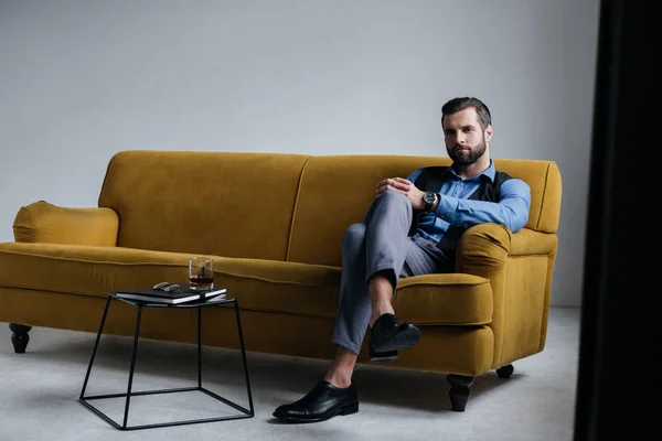 Modischer Mann sitzt auf gelbem Sofa mit Alkoholgetränk auf Tisch in der Nähe — Stockfoto