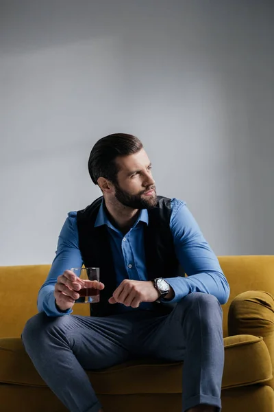 Hombre elegante sosteniendo bebida alcohólica y sentado en un sofá amarillo - foto de stock