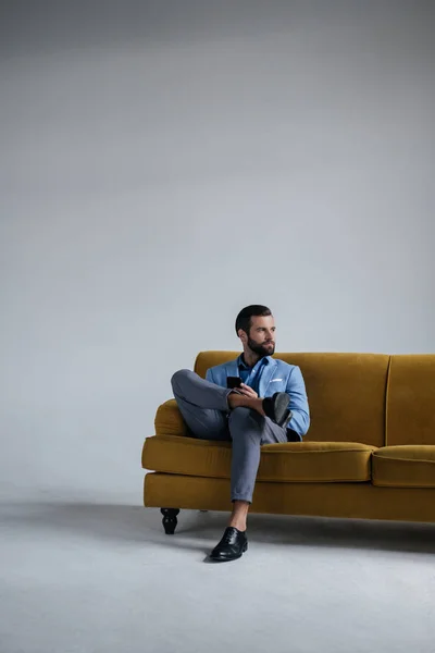 Élégant homme en costume à la mode en utilisant smartphone et assis sur un canapé jaune — Photo de stock