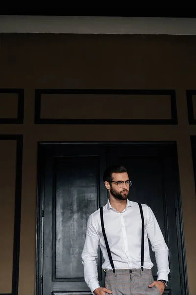 Handsome bearded man posing in white shirt and suspenders against door — Stock Photo