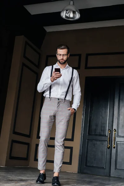 Elegante uomo barbuto in bretelle utilizzando smartphone in loft interno — Foto stock