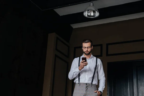 Homem barbudo elegante em suspensórios usando smartphone — Fotografia de Stock