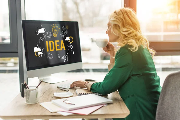 Middle aged businesswoman using desktop computer with idea icons on screen and drinking coffee at workplace — Stock Photo