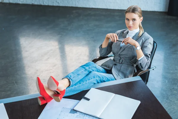 Hochwinkelaufnahme einer Geschäftsfrau, die mit Beinen auf einem Tisch im Büro sitzt und in die Kamera schaut — Stockfoto