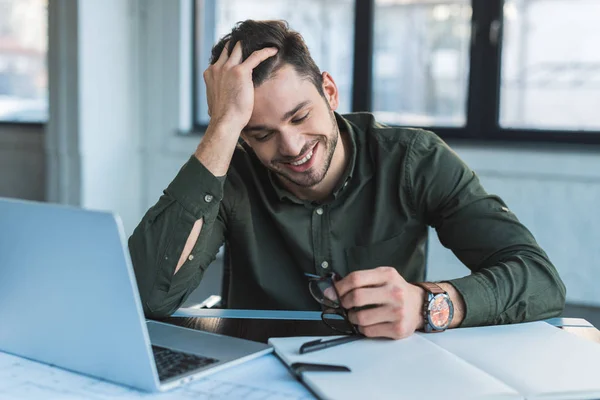 Uomo d'affari sorridente seduto a tavola in ufficio e con gli occhiali in mano — Foto stock