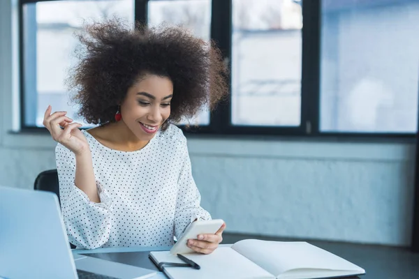 Donna d'affari afroamericana sorridente utilizzando smartphone in ufficio — Foto stock