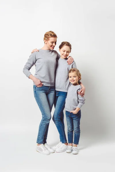 Mutter und Töchter verschiedener Generationen in ähnlicher Kleidung umarmen sich isoliert auf grau — Stockfoto