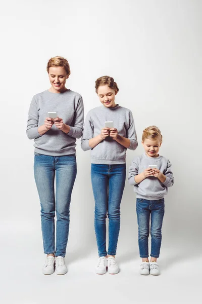 Madre e hijas en ropa similar utilizando teléfonos inteligentes aislados en gris - foto de stock