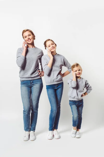 Madre e hijas en ropa similar hablando en teléfonos inteligentes aislados en gris - foto de stock