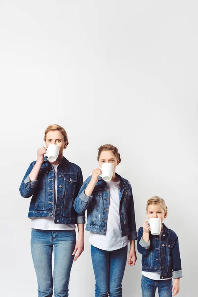 Портрет матери и дочери в похожей джинсовой одежде, пьющие горячие напитки, изолированные на сером — стоковое фото