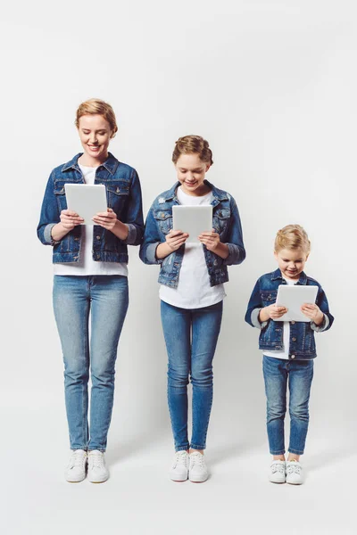 Lächelnde Mutter und Töchter in ähnlicher Jeanskleidung, die in einer Reihe stehen und Tabletten isoliert auf grau verwenden — Stockfoto