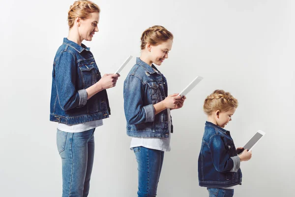 Мати і дочки в подібному джинсовому одязі, що стоїть в ряд і використовує таблетки ізольовані на сірому — стокове фото