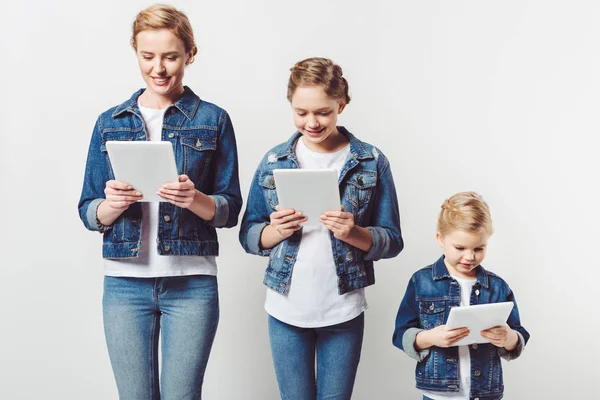 Mother and daughters in similar denim clothing standing in row and using tablets isolated on grey — Stock Photo