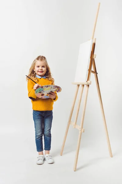 Adorable petit enfant avec pinceau et palette debout près de la toile sur chevalet sur blanc — Photo de stock