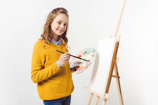 Happy teen female painter in yellow sweater with paint brush and palette near easel with blank canvas on white — Stock Photo