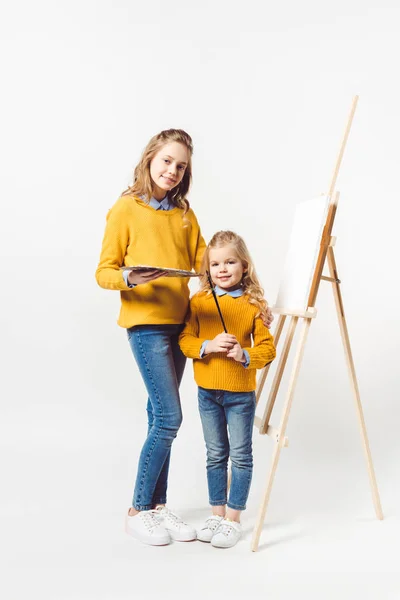 Sisters of different generations with palette and paint brush standing in front of easel with blank canvas — Stock Photo