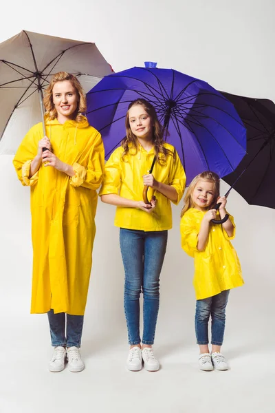 Happy mother and daughters in yellow raincoats with umbrellas standing in row on white — Stock Photo