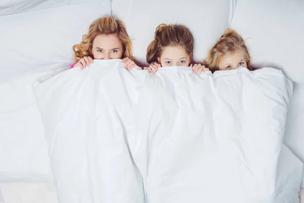 Vista superior de la madre y las hijas escondidas debajo de la manta en la cama - foto de stock
