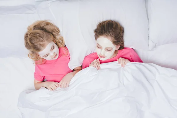 Top view of sisters with skincare facial masks lying in bed — Stock Photo