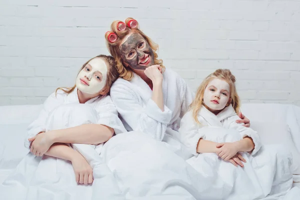 Madre e hijas en la cama con máscaras faciales de belleza - foto de stock