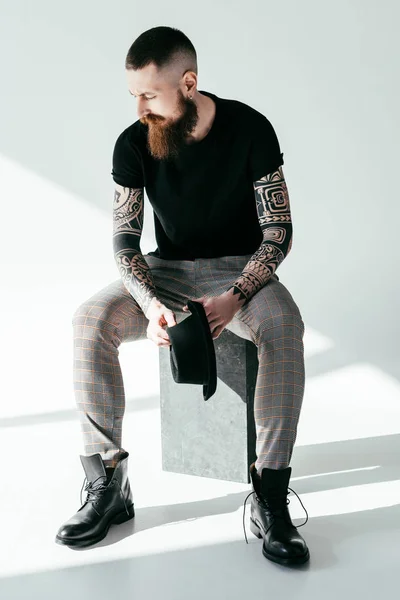 Élégant homme tatoué barbu assis et tenant chapeau sur blanc — Photo de stock