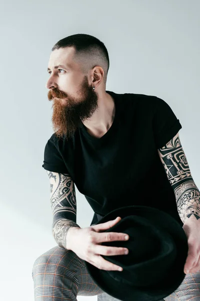 Handsome bearded tattooed man holding hat and looking away on white — Stock Photo