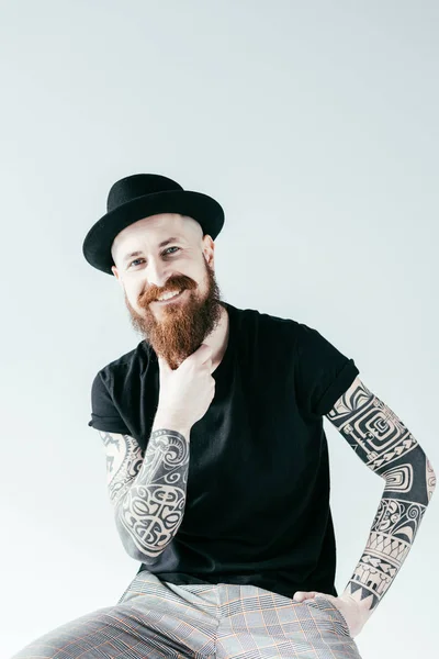 Sonriente hombre tatuado tocando la barba y mirando a la cámara aislada en blanco - foto de stock