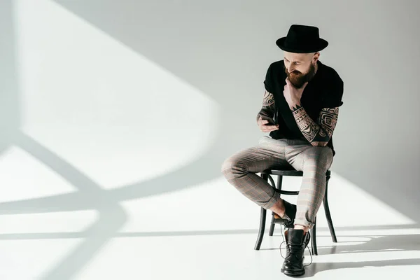 Красивый бородатый татуированный мужчина сидит на стуле и смотрит на смартфон на белом — стоковое фото