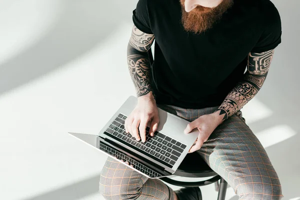 Cropped image of bearded tattooed man sitting on chair with laptop on white — Stock Photo