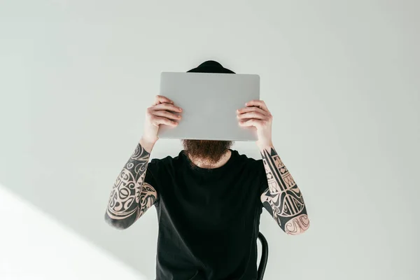 Bärtiger tätowierter Mann verdeckt Gesicht mit Laptop auf weißem Hintergrund — Stockfoto