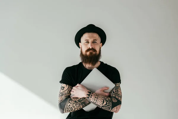 Красивый бородатый татуированный мужчина обнимает ноутбук и смотрит в камеру на белом — стоковое фото