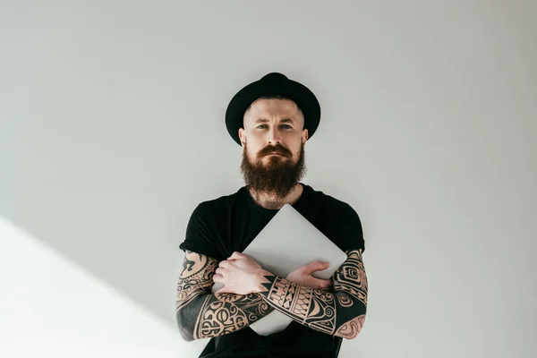 Bonito barbudo tatuado homem abraçando laptop e olhando para câmera no branco — Fotografia de Stock