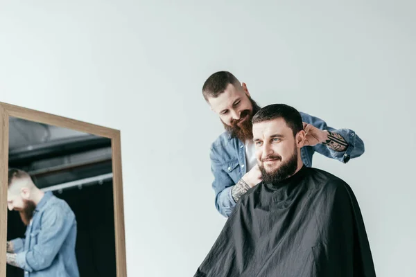 Barbería sonriente afeitar el pelo del cliente en la barbería aislado en blanco - foto de stock
