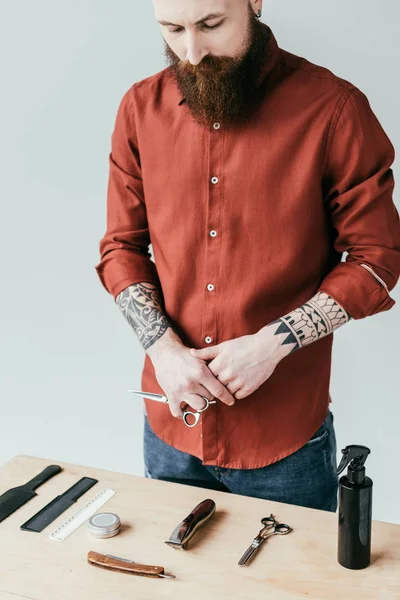 Bärtiger, gutaussehender Friseur, der eine Schere in der Hand hält und die Ausrüstung isoliert auf weißem Grund betrachtet — Stockfoto