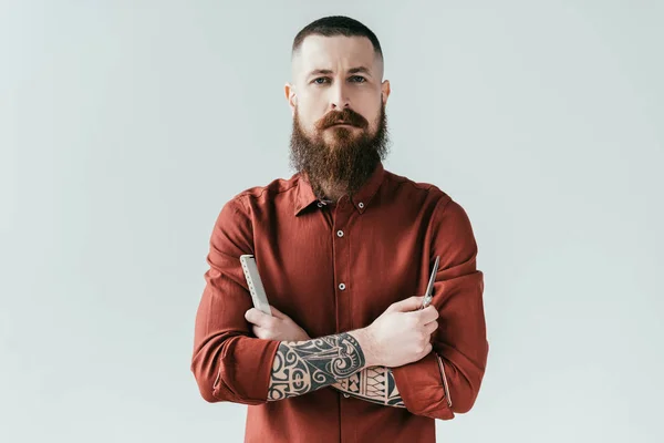 Barbero guapo barbudo sosteniendo peine y tijeras y mirando a la cámara aislada en blanco - foto de stock