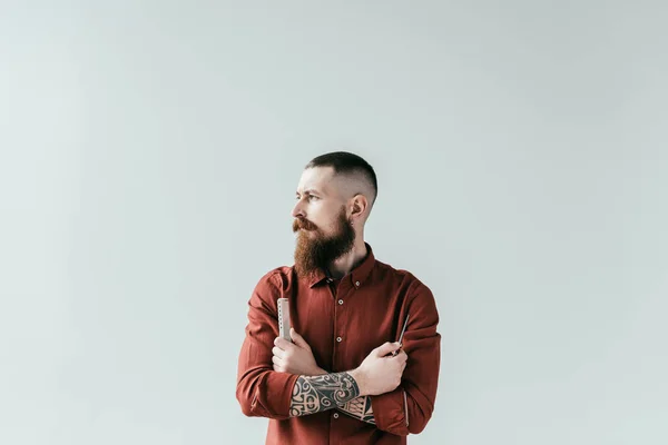 Barbero guapo barbudo sosteniendo peine y tijeras en brazos cruzados aislados en blanco - foto de stock