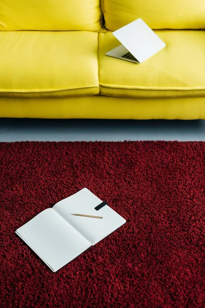 Half-closed laptop on yellow couch and textbook with pencil on rug — Stock Photo