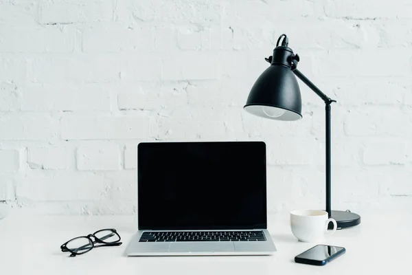 Laptop mit leerem Bildschirm, Smartphone, Lampe und Brille am Schreibtisch — Stockfoto