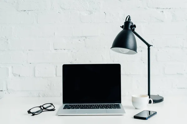 Laptop with blank screen, smartphone, lamp and eyeglasses on desk — Stock Photo