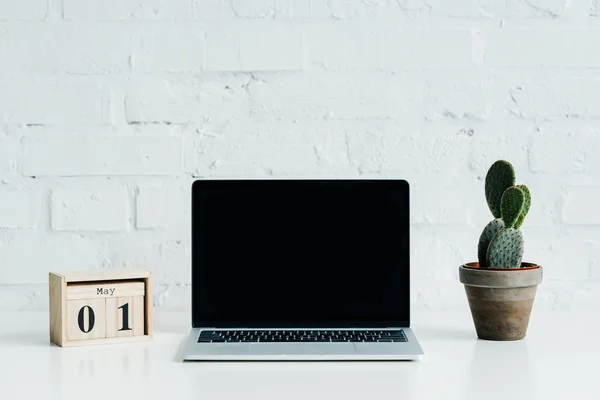 Calendario in legno con data 1 maggio, laptop con schermo bianco e impianto di cactus — Foto stock