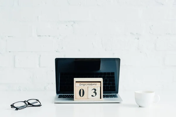 Calendário de madeira no laptop com tela em branco, óculos e xícara na superfície branca — Fotografia de Stock