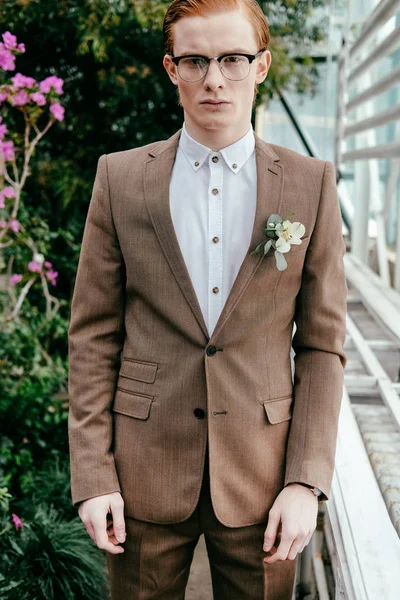 Retrato de hombre elegante con el pelo rojo en traje y gafas mirando a la cámara en invernadero - foto de stock