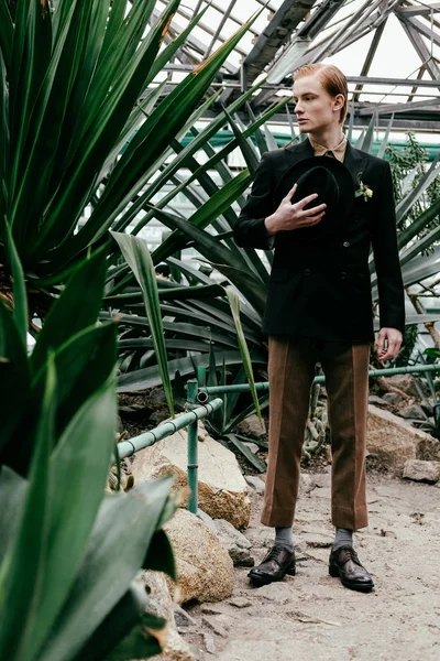 Élégant jeune homme aux cheveux roux avec chapeau à la main debout dans la serre — Photo de stock