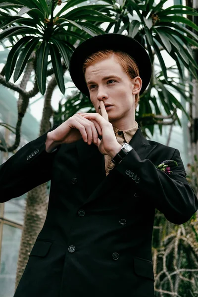 Portrait of stylish young red hair man in hat showing silence sign in glasshouse — Stock Photo