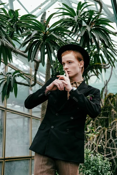 Portrait of stylish young red hair man in hat showing silence sign in glasshouse — Stock Photo