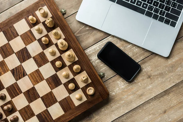 Smartphone, laptop y tablero de ajedrez con piezas de ajedrez en superficie de madera - foto de stock