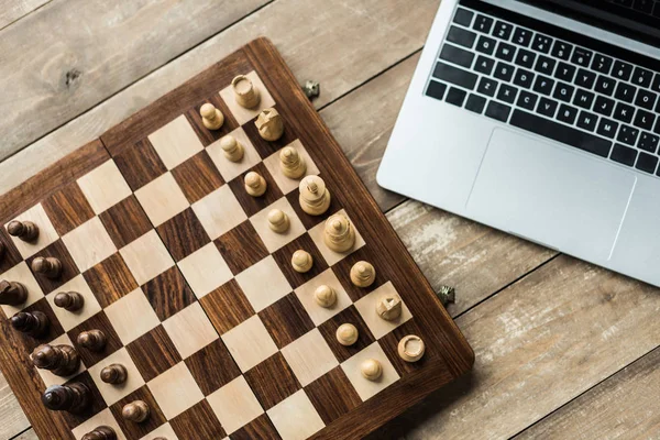 Laptop e tabuleiro de xadrez com figuras na superfície de madeira rústica — Fotografia de Stock