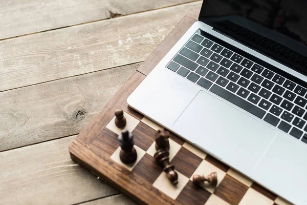 Chess board with chess pieces and laptop on rustic wooden surface — Stock Photo