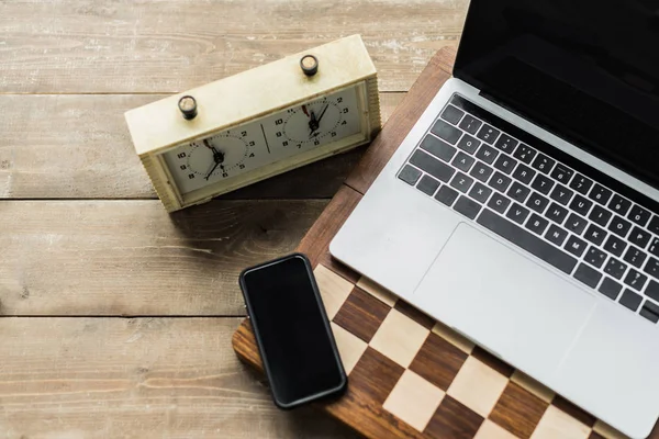 Orologio a scacchi, smartphone, laptop e scacchiera su superficie in legno — Foto stock