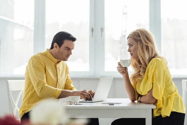 Blonde woman drinking coffee and looking at thoughtful man working on laptop — Stock Photo